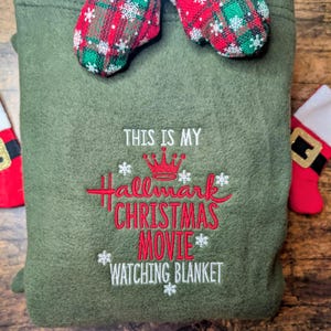 Puede incluir: Una manta verde oliva con el texto bordado "THIS IS MY Hallmark CHRISTMAS MOVIE WATCHING BLANKET" en rojo y blanco. La manta est&aacute; decorada con detalles de copos de nieve y rematada con mitones de cuadros rojos y verdes.