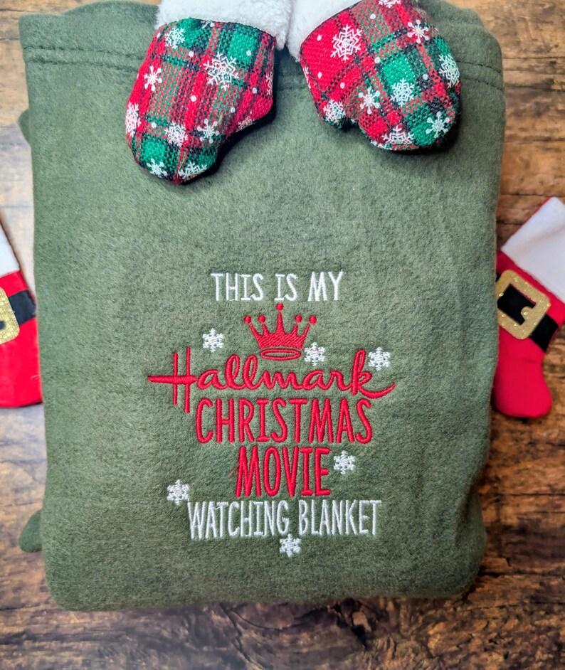 Puede incluir: Una manta verde oliva con el texto bordado "THIS IS MY Hallmark CHRISTMAS MOVIE WATCHING BLANKET" en rojo y blanco. La manta est&aacute; decorada con detalles de copos de nieve y rematada con mitones de cuadros rojos y verdes.