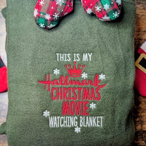 Puede incluir: Una manta verde oliva con el texto bordado "THIS IS MY Hallmark CHRISTMAS MOVIE WATCHING BLANKET" en rojo y blanco. Dos mini-mitones con patrones de cuadros rojos y verdes y ribete blanco est&aacute;n unidos en la parte superior.