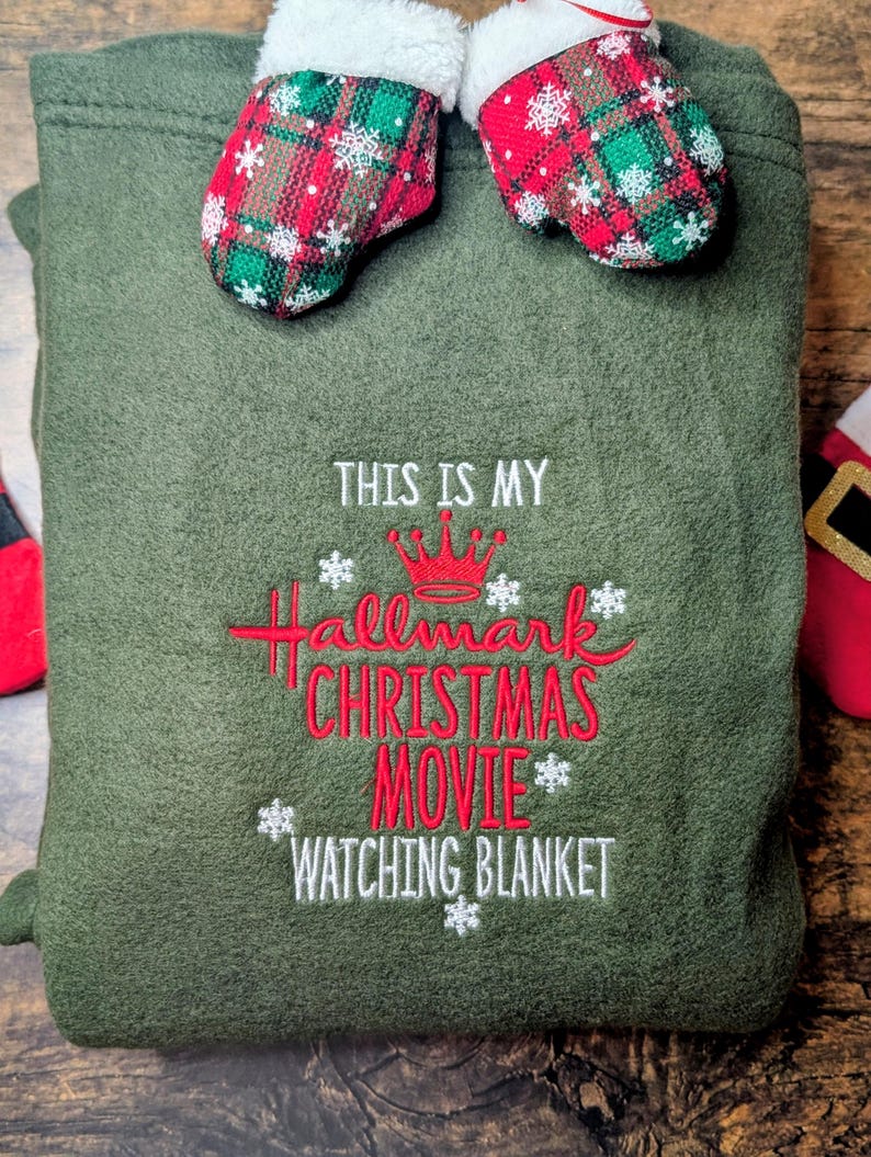 Puede incluir: Una manta verde oliva con el texto bordado "THIS IS MY Hallmark CHRISTMAS MOVIE WATCHING BLANKET" en rojo y blanco. Dos mini-mitones con patrones de cuadros rojos y verdes y ribete blanco est&aacute;n unidos en la parte superior.