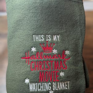 Puede incluir: Manta verde oliva doblada bordada con el texto "THIS IS MY Hallmark CHRISTMAS MOVIE WATCHING BLANKET" en rojo y blanco. Una corona roja y acentos de copo de nieve tambi&eacute;n est&aacute;n bordados en la manta.