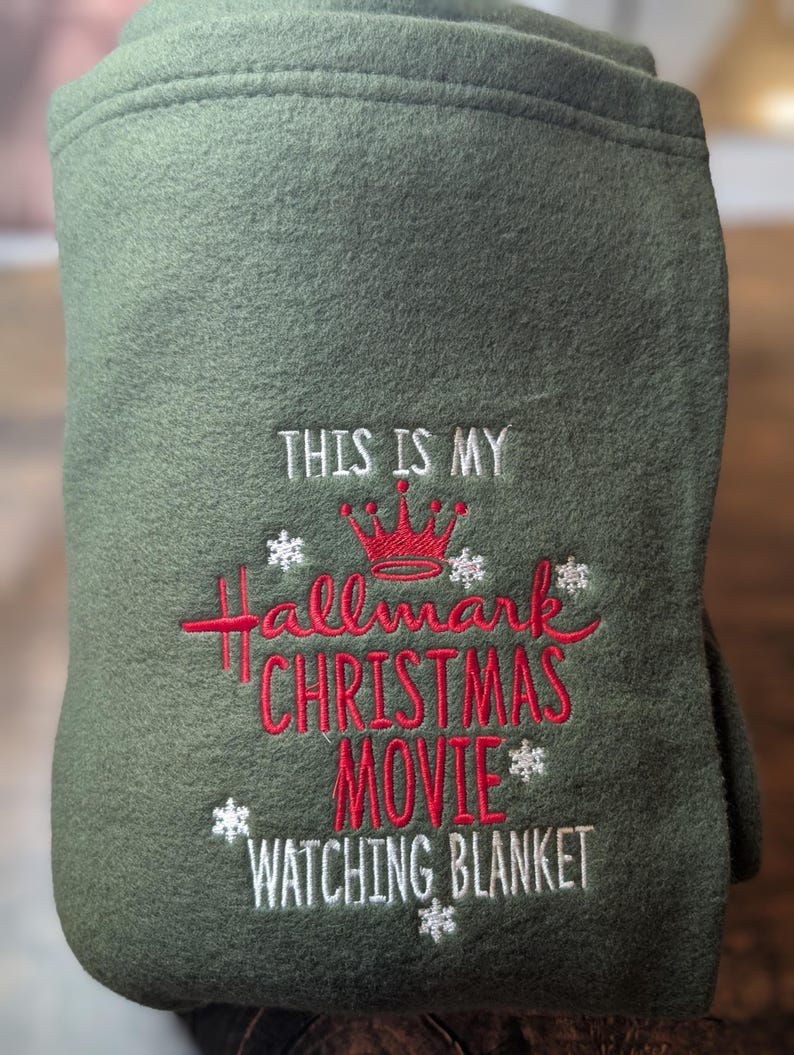 Puede incluir: Manta verde oliva doblada bordada con el texto "THIS IS MY Hallmark CHRISTMAS MOVIE WATCHING BLANKET" en rojo y blanco. Una corona roja y acentos de copo de nieve tambi&eacute;n est&aacute;n bordados en la manta.