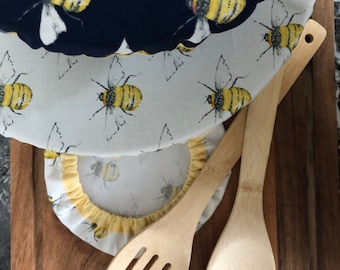 May include: Two wooden kitchen utensils, a slotted spoon and a serving spoon, on a wooden cutting board. The cutting board is covered with two fabric bee-themed bowl covers, one navy blue and one white with yellow bees.