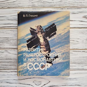 Può includere: Un libro vintage a tema spaziale. La copertina presenta un'illustrazione dettagliata di un satellite nello spazio su uno sfondo blu e bianco. Il titolo è in russo, con il nome dell'autore in alto.