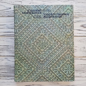 Puede incluir: Un libro de tapa dura con una cubierta estampada. La cubierta tiene un diseño geométrico repetitivo en tonos verdes y azules. El título está en un idioma extranjero. El libro está sobre una superficie de madera blanca.