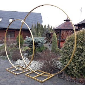 Könnte beinhalten: Zwei goldene Metallreifen, einer größer als der andere, stehen auf quadratischen Sockeln in einer Gartenlandschaft. Die Reifen sind nebeneinander angeordnet und bilden einen dekorativen Torbogen.