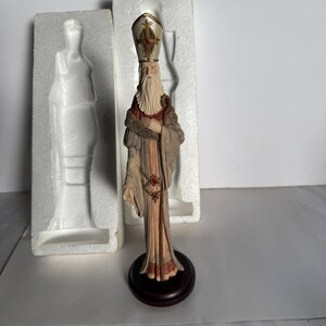 May include: A standing figurine of a bearded figure wearing a tall mitre hat, a long beige and brown robe with fur-like trim, and a decorative chain with a red gem. The figure is mounted on a circular dark wood base, positioned in front of white styrofoam.