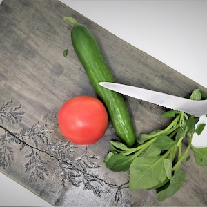 Puede incluir: Una tabla de cortar de madera con un acabado gris y un diseño de hojas. Un tomate rojo, un pepino verde y un manojo de hojas verdes están sobre la tabla. Un cuchillo dentado con mango rosa también está sobre la tabla.