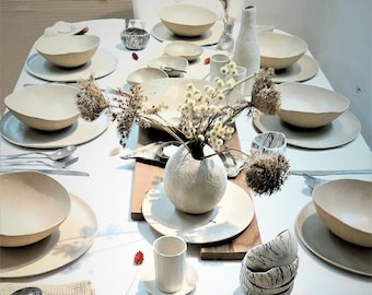 May include: A white table setting with a variety of ceramic bowls, plates, and cups. The table is decorated with a vase of white flowers and a wooden cutting board.