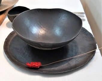 May include: A set of black ceramic bowls and plates on a white tablecloth. The bowls are deep and have a rounded shape. The plates are flat and have a slightly irregular shape. There are small red flowers on the table.