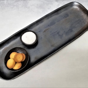 May include: A black rectangular serving tray with rounded edges, featuring two small bowls. One bowl contains white salt, and the other bowl holds several small, round, yellow cookies.