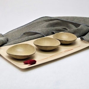 Puede incluir: Tres cuencos pequeños de cerámica marrón claro sobre una bandeja rectangular beige claro. La bandeja está sobre una superficie de tela gris.