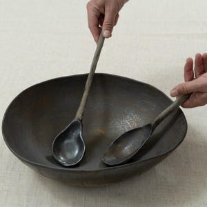 May include: A dark metal serving bowl with two matching spoons. The spoons have long, slender handles and rounded bowls. The bowl is wide and shallow, with a dark, textured surface. The spoons are held above the bowl.