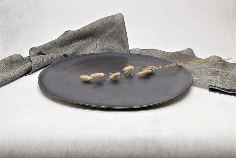 May include: A dark gray ceramic serving platter with a smooth, slightly textured surface. A sprig of dried, light brown grasses is placed on the platter.