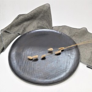 May include: A black ceramic serving platter with a textured surface. The platter is on a white surface with two gray linen napkins and a sprig of dried flowers.