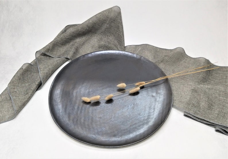 May include: A black ceramic serving platter with a textured surface. The platter is on a white surface with two gray linen napkins and a sprig of dried flowers.
