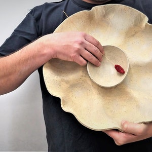 May include: A large, off-white ceramic bowl with a wavy, ruffled edge. A smaller, round bowl sits inside the larger bowl. A small, red object is in the smaller bowl.