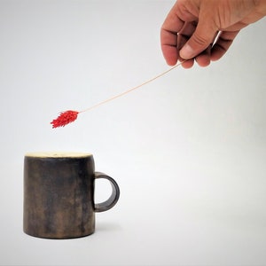 May include: A dark brown ceramic mug with a handle, sitting on a white surface. A single red flower is held by a hand, with the stem extending towards the mug.