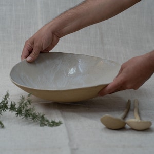 Può includere: Una grande ciotola in ceramica di forma ovale, con un interno bianco sporco macchiato e un esterno giallo chiaro. La ciotola è tenuta da due mani. Due piccoli cucchiai di legno sono visibili a destra della ciotola. Un rametto di verde è nell'angolo in basso a sinistra.