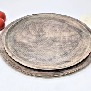 May include: Two wooden serving platters, one nested inside the other, with a rustic, gray-brown finish. The platters are on a white surface with red tomatoes and green herbs.