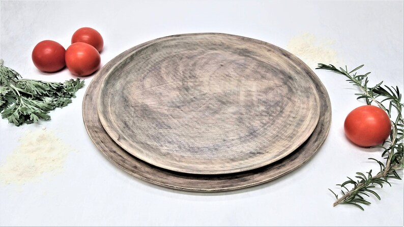 May include: Two wooden serving platters, one nested inside the other, with a rustic, gray-brown finish. The platters are on a white surface with red tomatoes and green herbs.