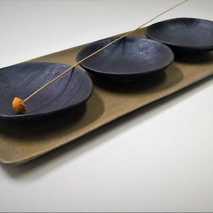 Puede incluir: Una bandeja rectangular con un glaseado marrón claro contiene tres cuencos pequeños negros. Un solo tallo de flor seca descansa sobre la bandeja.