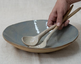 May include: A handmade ceramic serving bowl with a light blue interior and a tan rim. Two matching ceramic serving spoons are placed inside the bowl. The bowl and spoons have a rustic, speckled appearance, suggesting a natural, artisanal aesthetic.