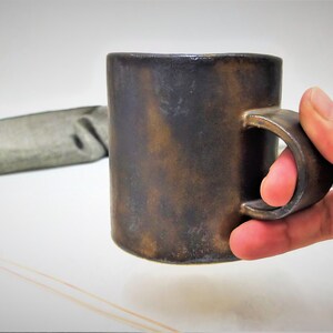 May include: A dark brown ceramic mug with a rounded handle. The mug has a rustic, aged look.