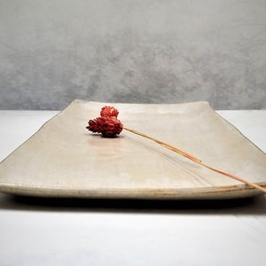 Pode incluir: Uma bandeja retangular de cerâmica com um esmalte bege claro e uma textura ligeiramente áspera. Uma única flor seca de cor castanho-avermelhada é colocada na bandeja.