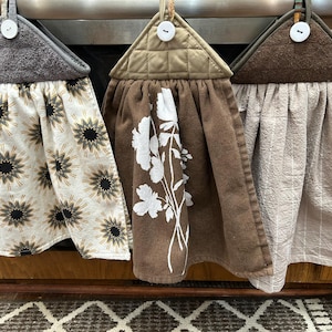 May include: Three hanging kitchen towels with decorative designs. One towel has a pattern of black and gold starbursts on a cream background. Another has white floral designs on a brown background. The third is a light brown striped towel.