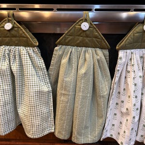 May include: Three hanging kitchen towels with quilted olive green tops and white buttons. The first towel features a green and white houndstooth pattern, the second is solid sage green with thin white stripes, and the third has a green floral print.