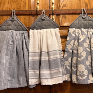 May include: Three hanging kitchen towels with gray quilted fabric tops and button closures. The left towel is light gray with white vertical stripes, the center is white with gray horizontal stripes, and the right is light gray with a white floral pattern.