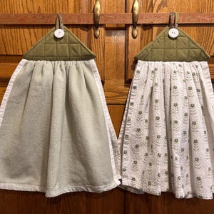 May include: Two hanging kitchen towels with olive green quilted fabric tops and white buttons. One towel is solid light sage green, while the other is white with a repeating small green floral pattern. Both feature a white border trim.