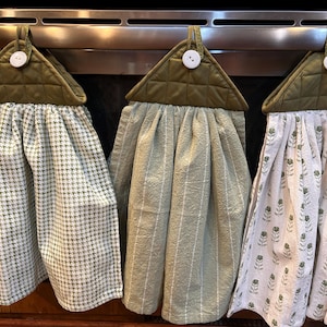 May include: Three hanging kitchen towels with olive green quilted fabric tops, each secured by a white button. The towels feature different patterns: a green and white houndstooth, a solid sage green with thin white vertical stripes, and a white floral print.