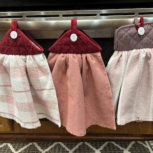May include: Three hanging kitchen towels in shades of red and pink. Each towel has a quilted top with a button detail and a loop for hanging. The towels have different patterns, including stripes and a waffle weave.