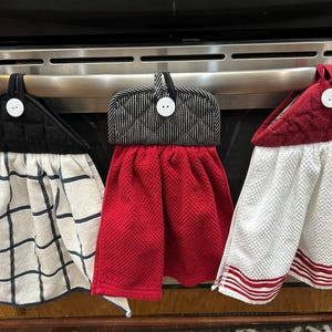 May include: Three kitchen hand towels hanging from a stainless steel oven handle. The towels have different designs: black and white plaid, red waffle weave, and white with red stripes. Each towel has a button.