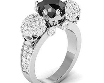 May include: A silver ring with a black gemstone surrounded by smaller white gemstones. The ring features two skull-shaped accents on either side of the gemstone, each adorned with white gemstones.