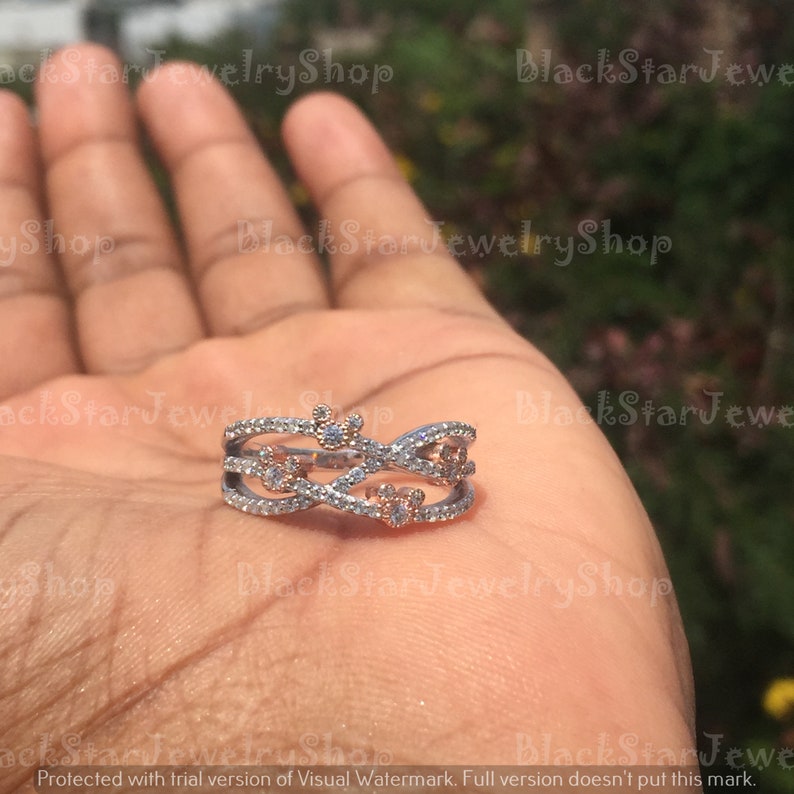 May include: A silver and rose gold ring with a delicate design featuring intertwined bands and small clear and pink stones. The ring has a Mickey Mouse silhouette in the center.