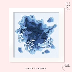 Peut inclure: Une illustration d'art découpé en papier bleu et blanc représentant une scène sous-marine avec un navire coulé, des poissons, des coraux et un crabe. L'œuvre est encadrée dans un cadre blanc et porte le texte "IDEAAVENUE" en bas.