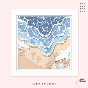 May include: A layered paper art piece depicting a beach scene. The artwork features blue ocean waves, a sandy shore with footprints, and white seagulls. The text "IDEA AVENUE" is at the bottom. The artwork is framed in a white square.