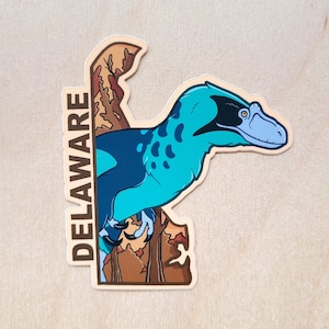 May include: A sticker featuring a cartoon dinosaur in shades of teal and blue, with the word "DELAWARE" printed vertically. The dinosaur is partially obscured by a brown and tan tree-like shape. The sticker is on a light wood grain surface.
