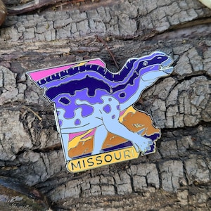 May include: Purple and blue dinosaur shaped enamel pin with a yellow outline. The pin is shaped like the state of Missouri and says "MISSOURI" in yellow letters at the bottom.