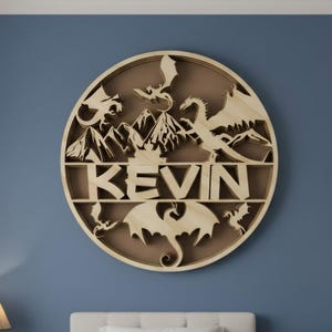 May include: A circular wooden wall art piece featuring a laser-cut design of mountains and multiple dragons surrounding the name KEVIN in bold, capital letters. The natural wood finish contrasts with the dark blue wall behind the display.