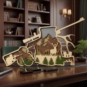 Könnte beinhalten: Ein mehrschichtiges, lasergeschnittenes Holzdisplay in Form eines Gewehrs, das eine Berglandschaft, einen Jäger, einen Hirsch und Tannenbäume zeigt. Das Design umfasst einen integrierten Stiftehalter und ein dekoratives Geweih-Element.