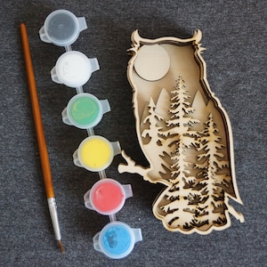 Op de afbeelding: Een houten uil-uitsparing met een maan en dennenbomen erin. De uil is klaar om te worden geschilderd met zes kleine potjes verf: zwart, wit, groen, geel, rood en blauw. Een kwast is inbegrepen.