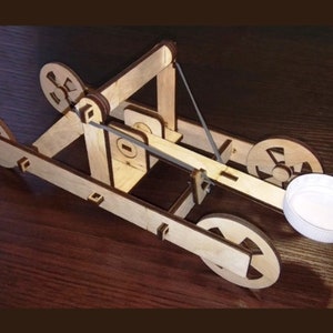 May include: A wooden toy catapult with a white plastic lid in the launching arm. The catapult has a simple design with wooden wheels and a lever.