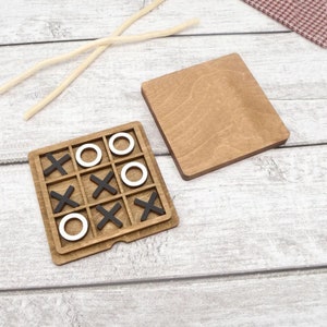 May include: A wooden tic-tac-toe game with black X's and white O's. The game is set up on a wooden board with a separate wooden lid.