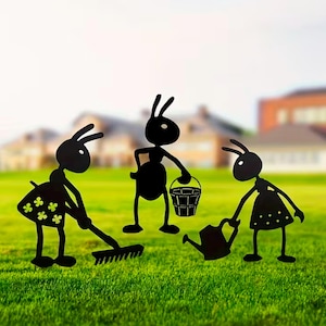 May include: Black metal garden ornaments depicting three ants. One is raking, another carries a bucket, and the third waters plants. The ants are on a green lawn with a blurred background of houses and sky.
