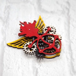 May include: A wooden steampunk-style brooch with a red flame design, yellow wings, and intricate gears.