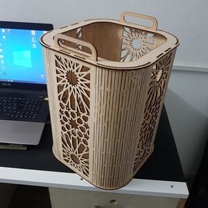 Puede incluir: Una cesta de madera de color claro con un patrón geométrico de estrella cortado en los lados. La cesta tiene dos asas y está sobre una superficie negra. El diseño es intrincado y decorativo.